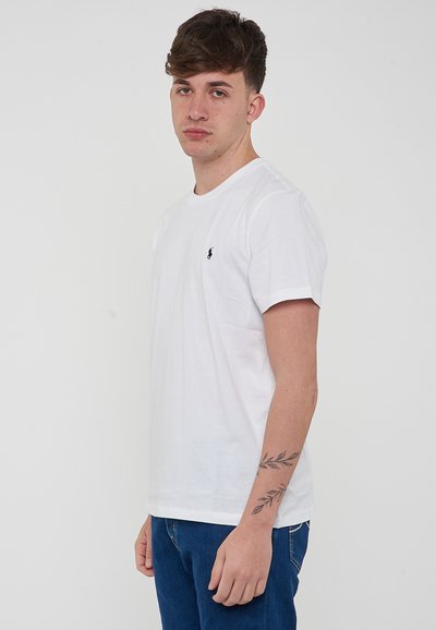 T-shirt bianco in cotone con maniche corte, colletto rotondo e un piccolo logo blu navy sul lato sinistro del petto, abbinato a jeans in denim blu.