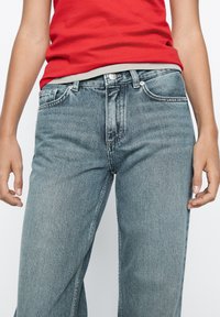 Hellblaue Denim-Jeans mit geradem Schnitt, sichtbaren Nähten, Knopf- und Reißverschluss, getragen mit einem roten Tanktop.