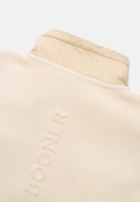 Cremefarvet fleece-pullover med en glat, struktureret overflade. Har en kontrasterende lys beige krave og broderet "BOGNER" detalje.