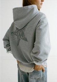 Sweat à capuche gris avec un grand motif étoile sur le dos. Il présente des poignets côtelés et un look superposé avec une chemise plus claire en dessous.