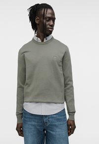 Χακί πλεκτό πουλόβερ με στρογγυλεμένο λαιμόκοψη και διακριτικό λογότυπο, φορεμένο πάνω από ανοιχτό γκρι πουκάμισο με γιακά και συνδυασμένο με μπλε τζιν.