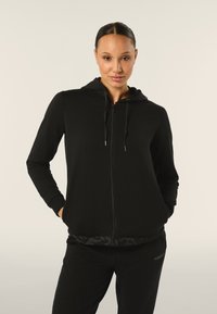 Felpa nera con zip, dal tessuto morbido, con cappuccio regolabile e tasche laterali. Abbinata a pantaloni neri coordinati con dettagli del logo.