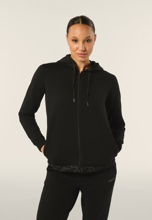 Haut à capuche zippé noir avec une texture douce, capuche ajustable à cordon, et poches latérales. Associé à un pantalon noir assorti avec un détail de logo.