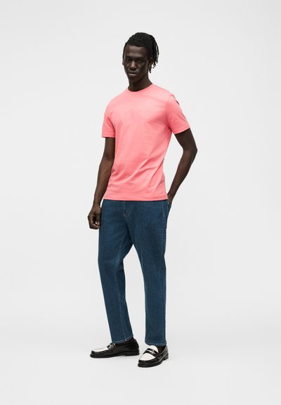 Homme à la peau foncée et dreadlocks portant un t-shirt rose vif à manches courtes, un jean bleu et des mocassins noirs et blancs, debout avec une main dans la poche.