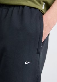 Schwarze Sporthose mit elastischem Bund, ausgestattet mit einer Seitentasche und einem weißen Nike-Logo, das auf dem linken Oberschenkel gestickt ist.