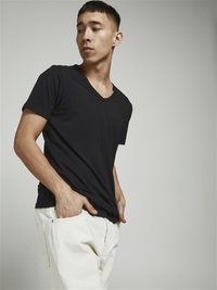 T-shirt noir à col en V dans une coupe décontractée, en coton doux. Associé à un pantalon texturé de couleur claire. Le mannequin pose les mains dans les poches.