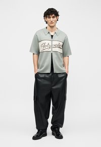 Jaunas vyras stovi dėvėdamas pilką trumpomis rankovėmis užsegamą su užtrauktuku marškinius su užrašu „Holzweiler“ ir laisvus juodus odinius kelnes su juodais batais.