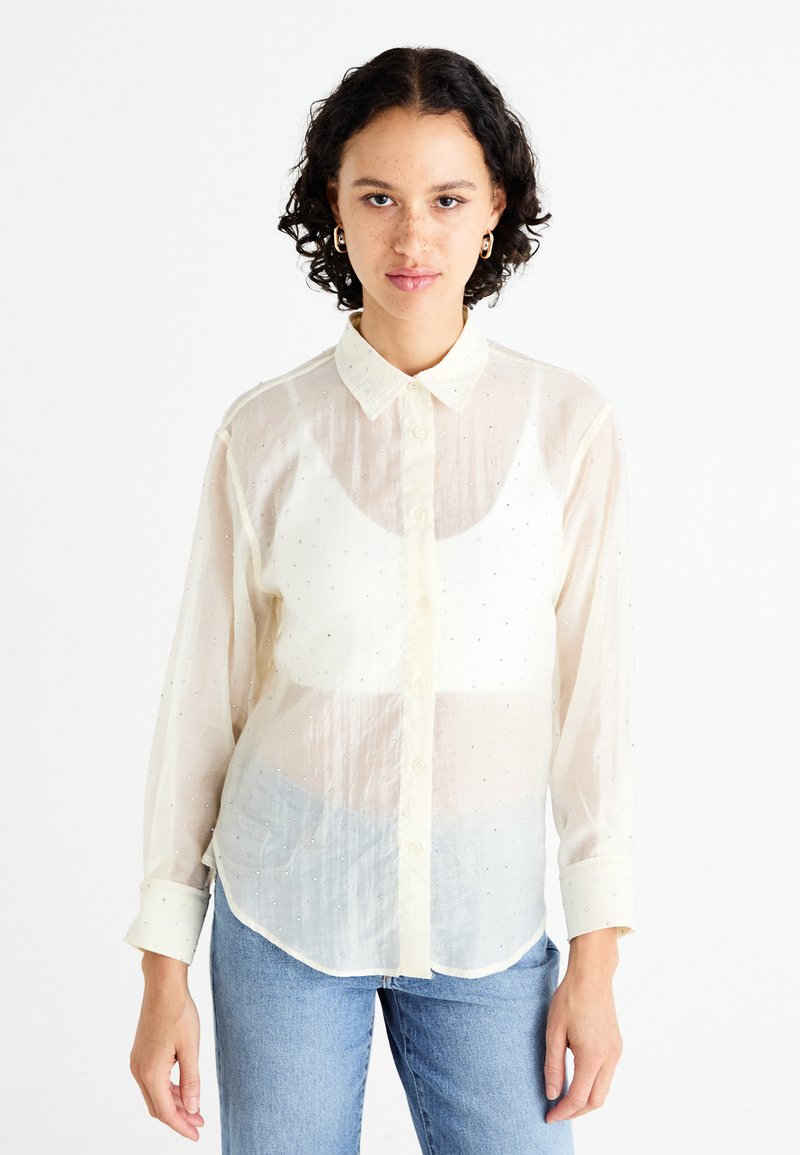 Camisa abotonada ligera y transparente en color blanco roto con una textura suave, que presenta pequeños adornos y un cuello clásico.