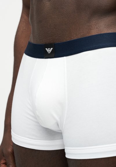 Κοντινό πλάνο ανδρικού κορμού με σκούρο χρώμα δέρματος, που φοράει λευκά μποξεράκια με μπλε σκούρο ελαστικό περιλαίμιο και ένα μικρό μαύρο εμβληματικό σημάδι στο κέντρο μπροστά.