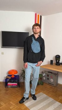 Svjetloplavi crossbody torba od tkanine s glatkom teksturom, nošena preko crne zip-up dukserice, u kombinaciji s svijetlim trapericama i crnim cipelama.