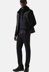 Man in een zwart leren jack met gebreide mouwen, marineblauw shirt, donkere jeans, zwarte sneakers en een zwarte pet, staand met één hand in zijn zak.