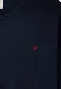 Suéter azul marino con escote redondo y textura acanalada. Presenta un pequeño logotipo de lazo rojo bordado en el lado izquierdo del pecho.