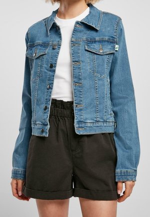 Persona con una chaqueta de mezclilla azul con cierres de botones sobre una camisa blanca y pantalones cortos negros de cintura alta enrollados.