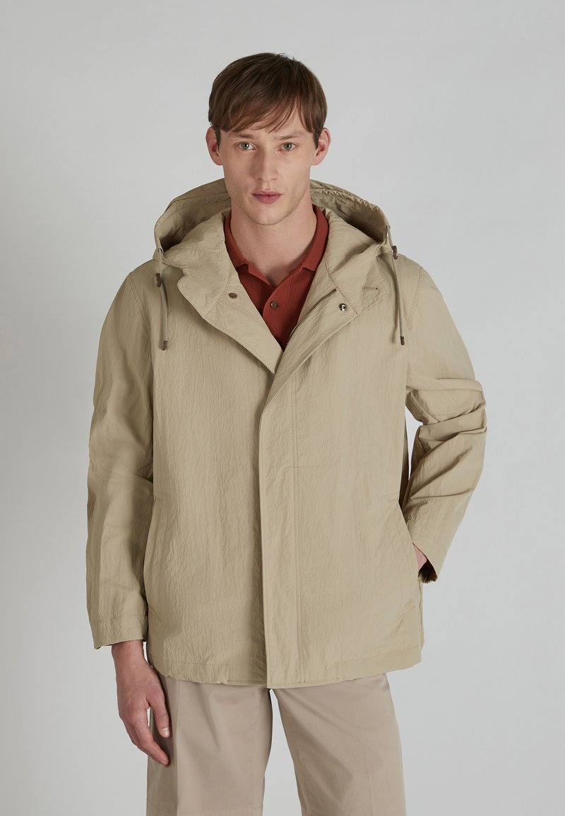 Chaqueta beige con capucha, de tela texturizada, con bolsillos laterales y cierre de botón, llevada sobre una camiseta polo color burdeos.