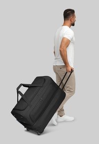 Borsa da viaggio nera con ruote, realizzata in tessuto liscio e testurizzato, con manici lunghi, manico telescopico e ruote tonde, trainata da una persona.