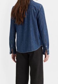 Donna con capelli lunghi e castani, che indossa una camicia di denim blu e pantaloni neri, vista da dietro su uno sfondo chiaro.
