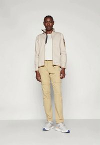 Világos bézs színű cipzáras dzseki magas gallérral, khaki nadrággal párosítva. Az öltözetet szürke és kék sportcipők egészítik ki, amelyek textúrával és rétegelt dizájnnal rendelkeznek.