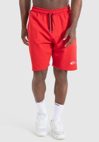 Rode sportieve shorts met een elastische tailleband en een zwarte trekkoord. Bevat een wit logo op de onderlinkerkant. Gemaakt met witte sneakers en sokken.