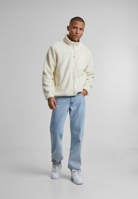 Giacca in pile color crema con colletto alto, chiusura con zip e taschino. Indossata con jeans azzurri e sneakers bianche.