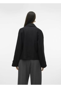 Giacca nera con un design su misura, caratterizzata da colletto e maniche lunghe. Il tessuto ha una texture liscia. Abbinata a pantaloni grigi.