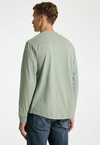 Cămașă lungă cu mâneci, de culoare verde deschis, cu guler rotund, textură netedă și cusături minime, purtată cu blugi negri cu un design clasic al buzunarelor.