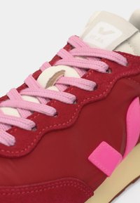 Κόκκινο και ροζ αθλητικό παπούτσι με ανοιχτό ροζ κορδόνια, μπεζ γλώσσα και φωτεινό ροζ λογότυπο στο πλάι, που εμφανίζει κοντινές λεπτομέρειες και ραφές.