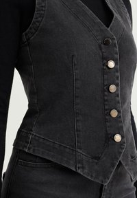 Svart denimväst med V-ringning, fem metallknappar och snedställda sömmar, med en figursydd silhuett och slät textur.