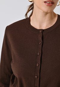 Cardigan marrone realizzato in morbido tessuto a maglia, con scollo rotondo, chiusure a bottone e una texture liscia.