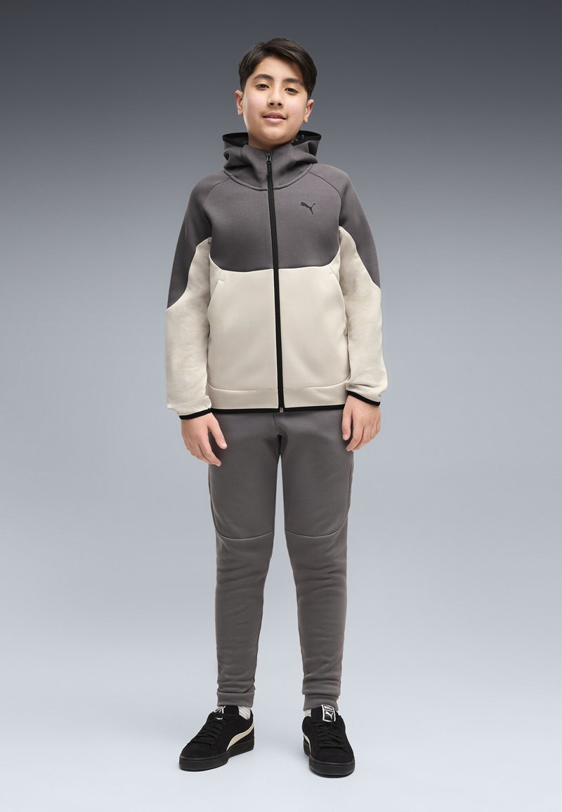 Garçon debout portant un sweat à capuche zippé bicolore gris et beige, un pantalon gris assorti et des baskets noires Puma sur un fond gris uni.