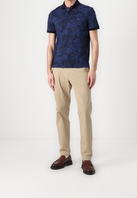 Granatowa koszulka polo w wzór palmowych liści, krótki rękaw i kołnierzyk, w połączeniu z beżowymi spodniami i brązowymi butami wsuwanymi.