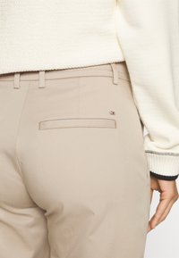 Beige broek met een gladde textuur, voorzien van een achterzak en een klein logo-accent. Gekleed met een crèmekleurige, getextureerde trui.