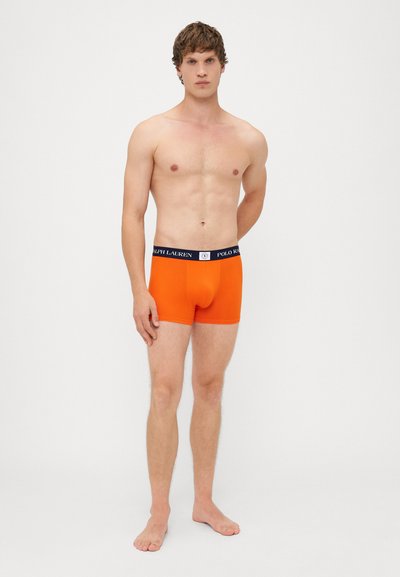 Oranžové boxerky s černým pasem, na kterém je bílým písmem nápis "RALPH LAUREN". Tkanina vypadá hladce a přiléhavě.