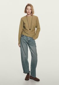 Frau steht mit den Händen in den Taschen, trägt einen olivgrünen Pullover, lockere blaue Jeans, braune Schuhe und eine lange Halskette mit einem runden Anhänger.
