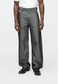 Platas džinsi tumši pelēkā džinsa audumā ar plašām kājām. Aprīkoti ar pogu un rāvējslēdzēja aizdari, priekšējām kabatām un gludu, teksturētu apdari. Valkāti ar melniem apaviem.