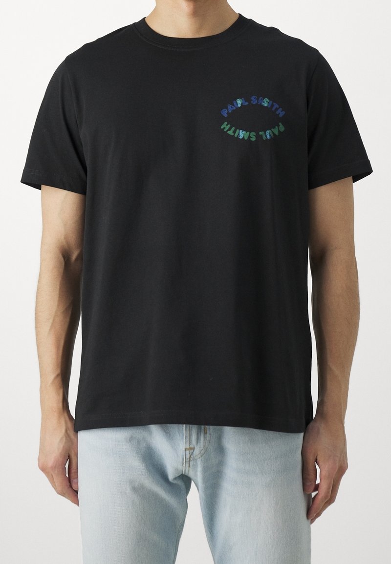 Homme portant un t-shirt noir à manches courtes avec le texte incurvé "PAUL SMITH" sur la poitrine gauche, associé à un jean bleu clair.