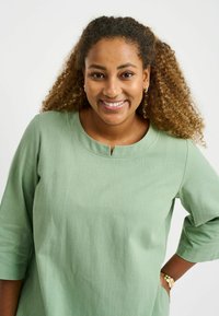 Green linen top with three-quarter sleeves, round neckline, and a small front opening detail. Smooth texture with a subtle weave pattern.