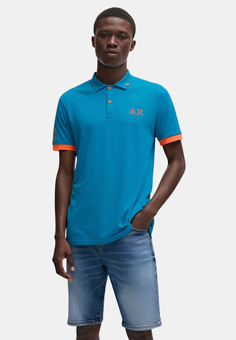 Polo shirt color turchese con accenti arancioni alle maniche, dotato di colletto e logo. Abbinato a pantaloni corti in denim, che mostrano una vestibilità rilassata e uno stile casual.