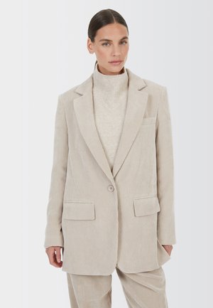 Frau trägt ein beiges Cord-Blazer und passende Hose über einem hellbeigen Rollkragenpullover, steht vor einem schlichten Hintergrund.