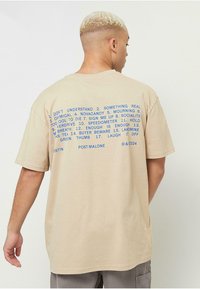 Smėlinis medvilninis marškinėliai su trumpomis rankovėmis, ant nugaros užrašyta mėlyna spalva, nurodant frazes ir "Post Malone" apačioje.
