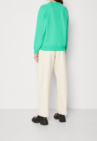Persona vestida con sudadera verde menta, pantalones anchos color blanco roto y zapatos de tacón grueso negros, de pie contra un fondo blanco liso.