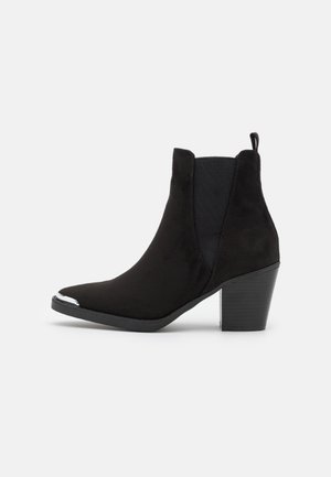 Bottine noire en cuir suédé, avec un bout pointu, des panneaux latéraux élastiques et un talon épais orné d'un accent métallique sur le bout.