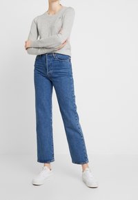 Hellgrauer Pullover, blaue hochgeschnittene Jeans und weiße Sneakers. Der Pullover hat einen runden Halsausschnitt; die Jeans haben gerade Beine und eine klassische Passform.