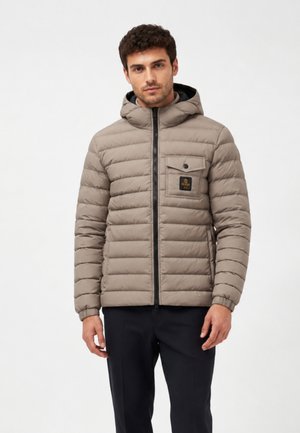Uomo con giacca imbottita con cappuccio marrone chiaro con zip frontale e tasca sul petto, abbinata a pantaloni neri su sfondo semplice.
