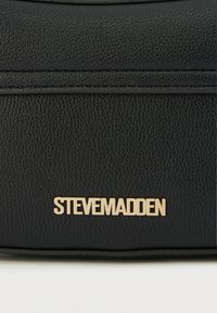 Geantă de mână din piele neagră, cu suprafață texturată, detalii cusute și un accent de logo "STEVE MADDEN" în ton auriu pe partea din față.