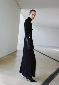 Robe noire à manches longues et col roulé avec une silhouette cintrée, associée à des gants en cuir noirs et des escarpins à bout pointu. Fond neutre.