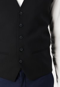 Gilet noir en tissu à la texture lisse, avec six boutons noirs et deux poches étroites à la taille. Coupe ajustée.