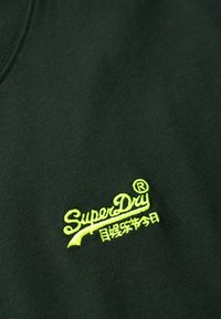 Tessuto verde scuro con una superficie liscia, caratterizzato da un logo ricamato giallo-verde brillante che riporta la scritta "SuperDry" in un carattere stilizzato.
