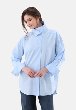 Camicia oversize azzurro chiaro in tessuto morbido, caratterizzata da un colletto alto, chiusura con bottoni e ampie maniche. Abbinata a pantaloni in denim scuro.