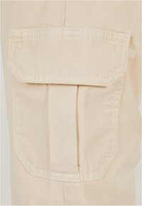 Detailní záběr béžové látkové kapsy s klopou a viditelným švem, připevněné k odpovídajícímu béžovému oděvu.