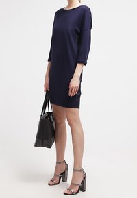 Abito a maglia blu navy con maniche a tre quarti, abbinato a sandali con tacco argentati e una borsa nera decorata con borchie.